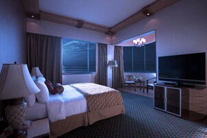 Habitación de hotel con cama, televisión, escritorio con lámpara, silla y ventana grande con cortinas.