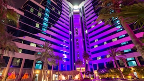 Un hotel de varios pisos con zona de piscina, palmeras y una fuente central.