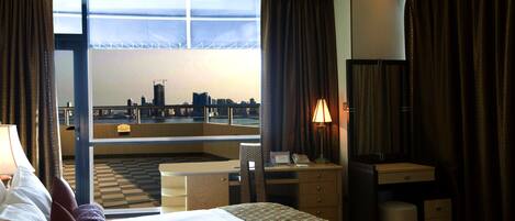 A hotel room with a bed, a desk, a chair, a lamp, and a view of a cityscape through large windows.