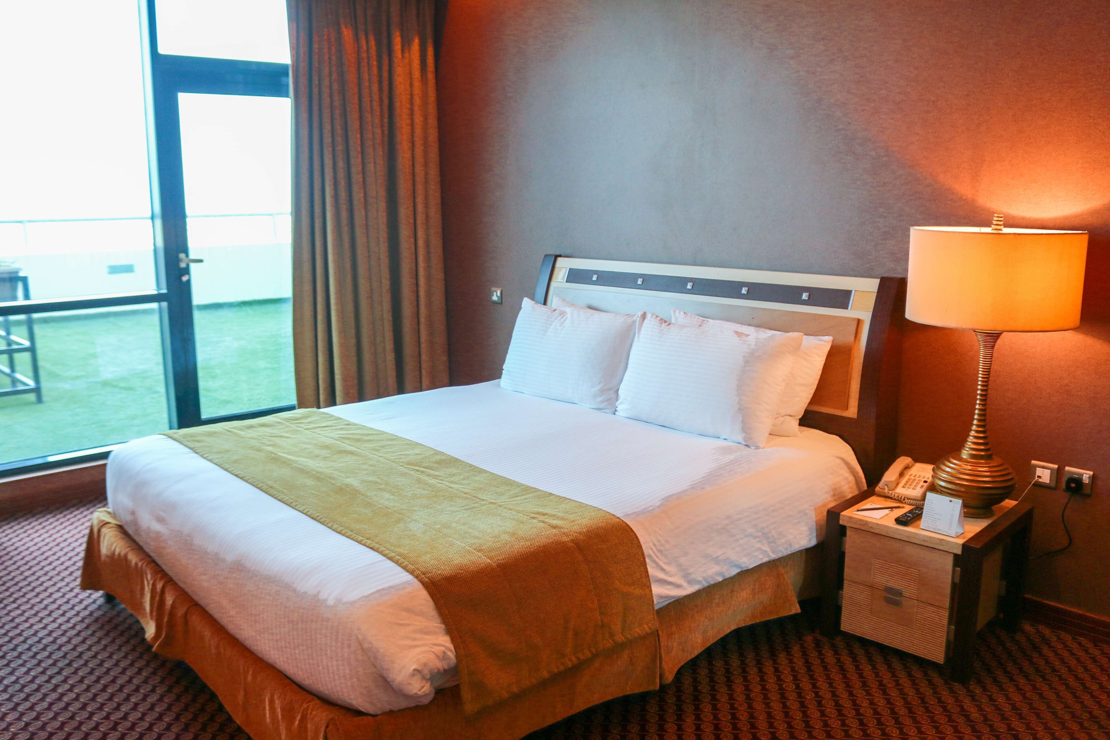 A hotel room with a large bed, a bedside lamp, a nightstand, and a view of the outdoors.