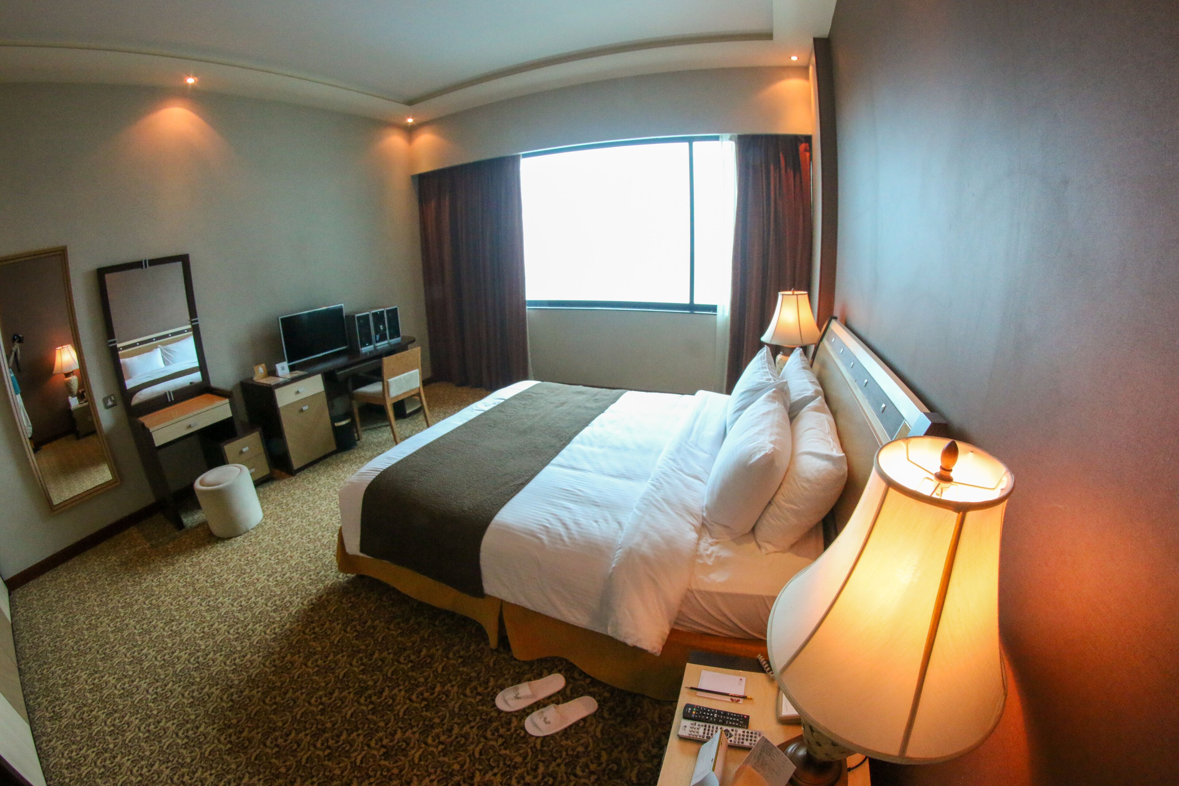 Habitación de hotel con una cama grande, un escritorio con computadora, televisor y ventana con cortinas.