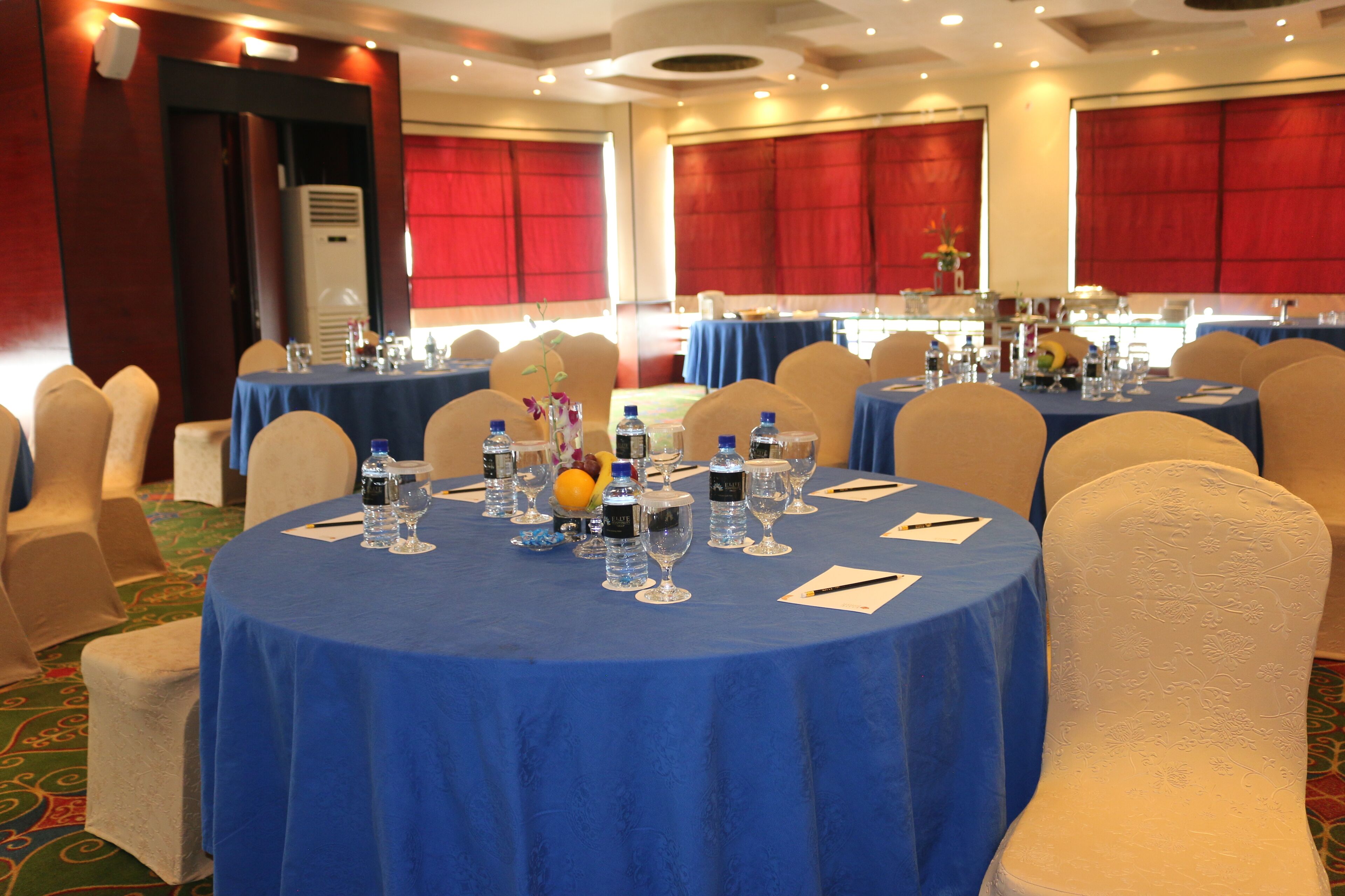Sala de conferências com mesas redondas cobertas por toalhas azuis, equipada com garrafas de água, copos e blocos de anotações.