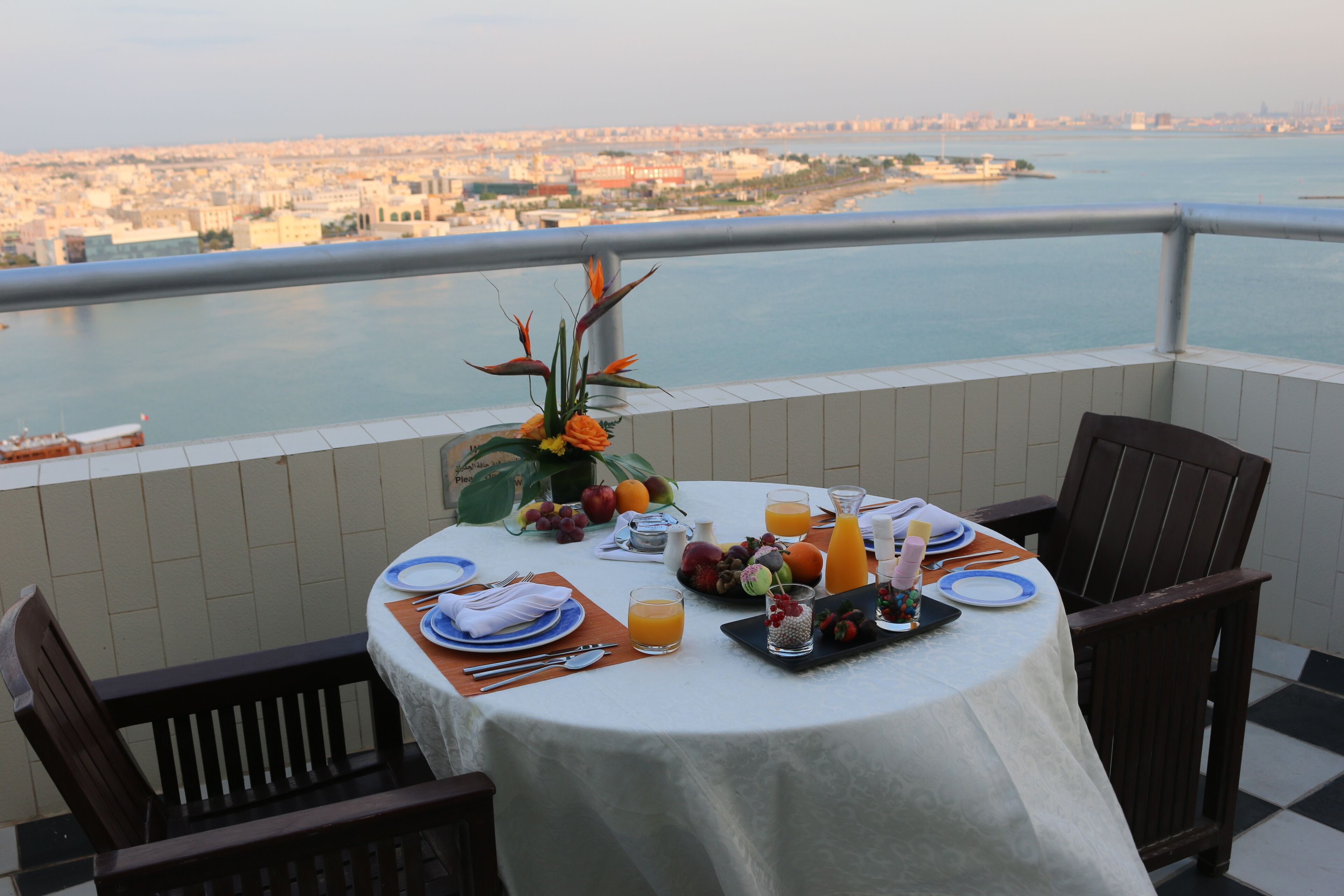 Une table ronde recouverte d’une nappe blanche, dressée pour le petit-déjeuner, avec vue sur un paysage urbain et une étendue d’eau.