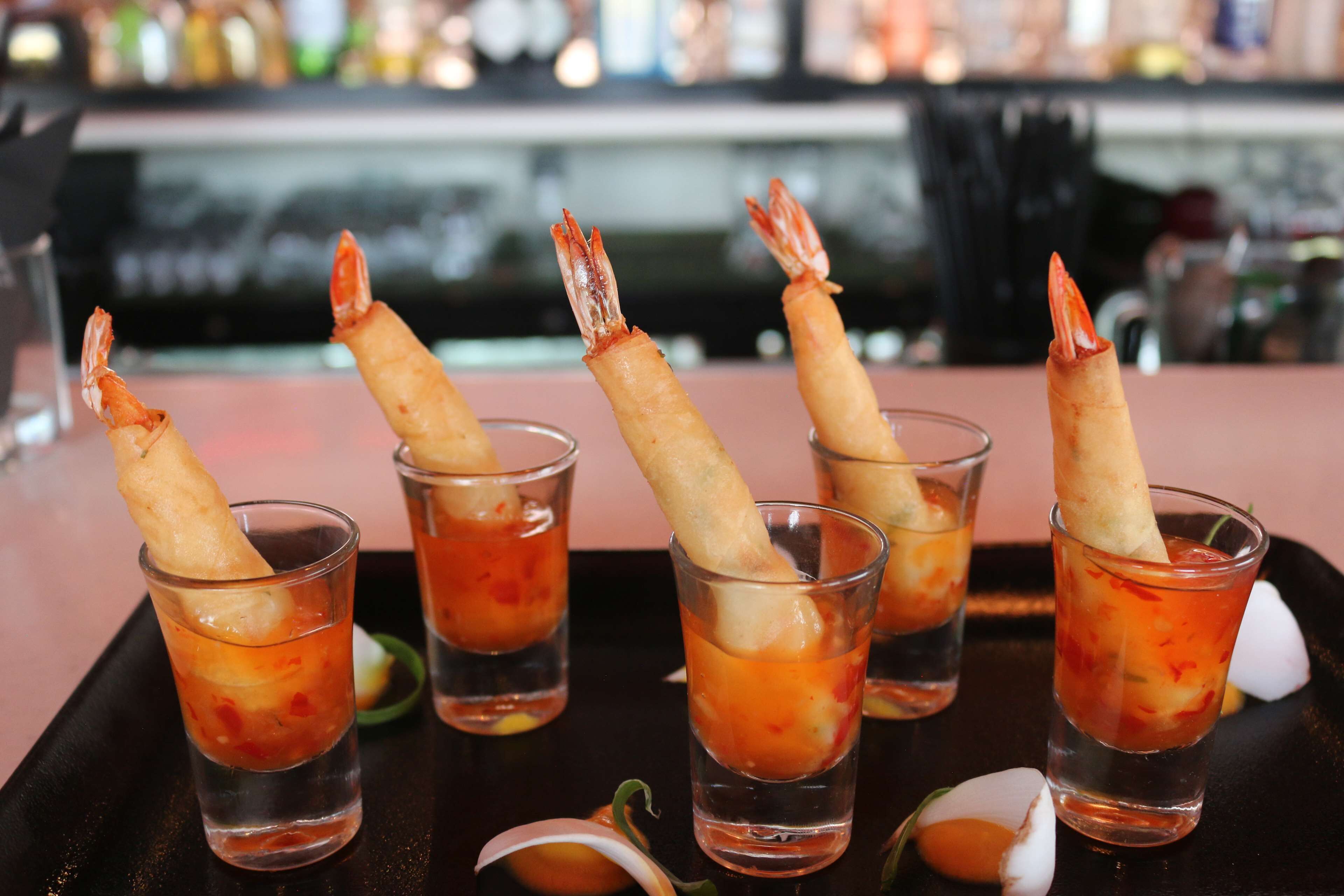 Una bandeja con cinco vasos para shots, cada uno conteniendo una bebida roja y adornado con un tempura de camarón en el borde.