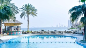 Una piscina con sillones de descanso y una palmera, un pequeño edificio con techo de paja, y un horizonte urbano al fondo.