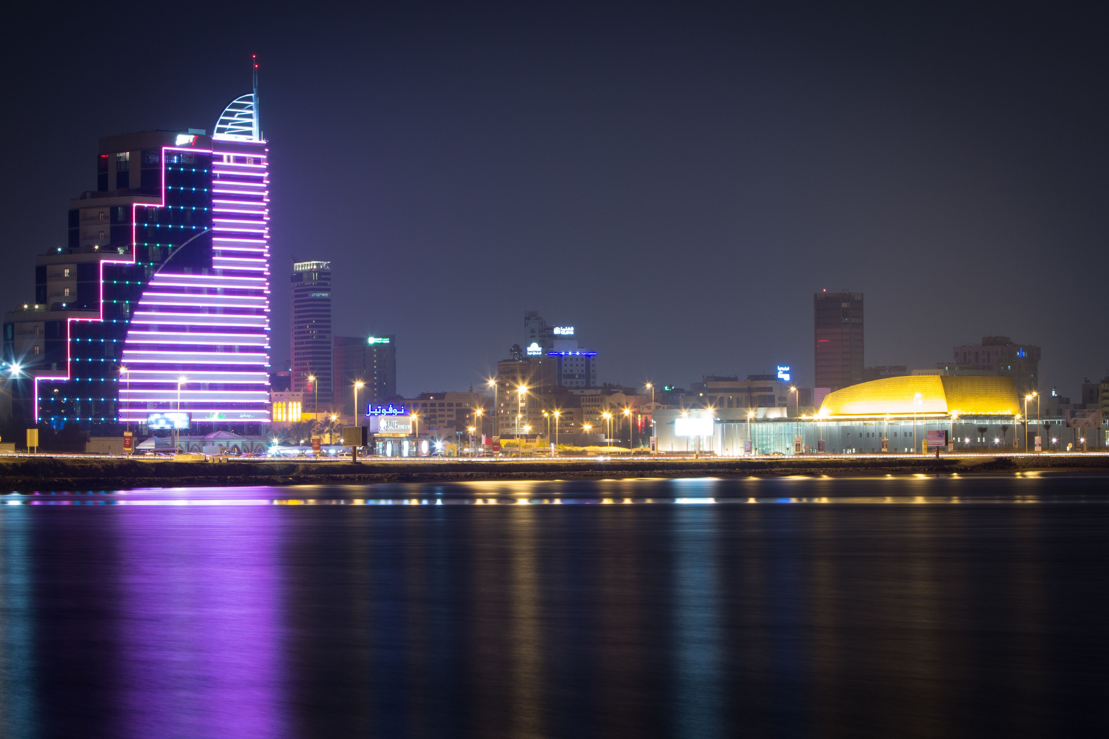 A nighttime cityscape with illuminated buildings, a river in the foreground, and a prominent yellow dome structure.