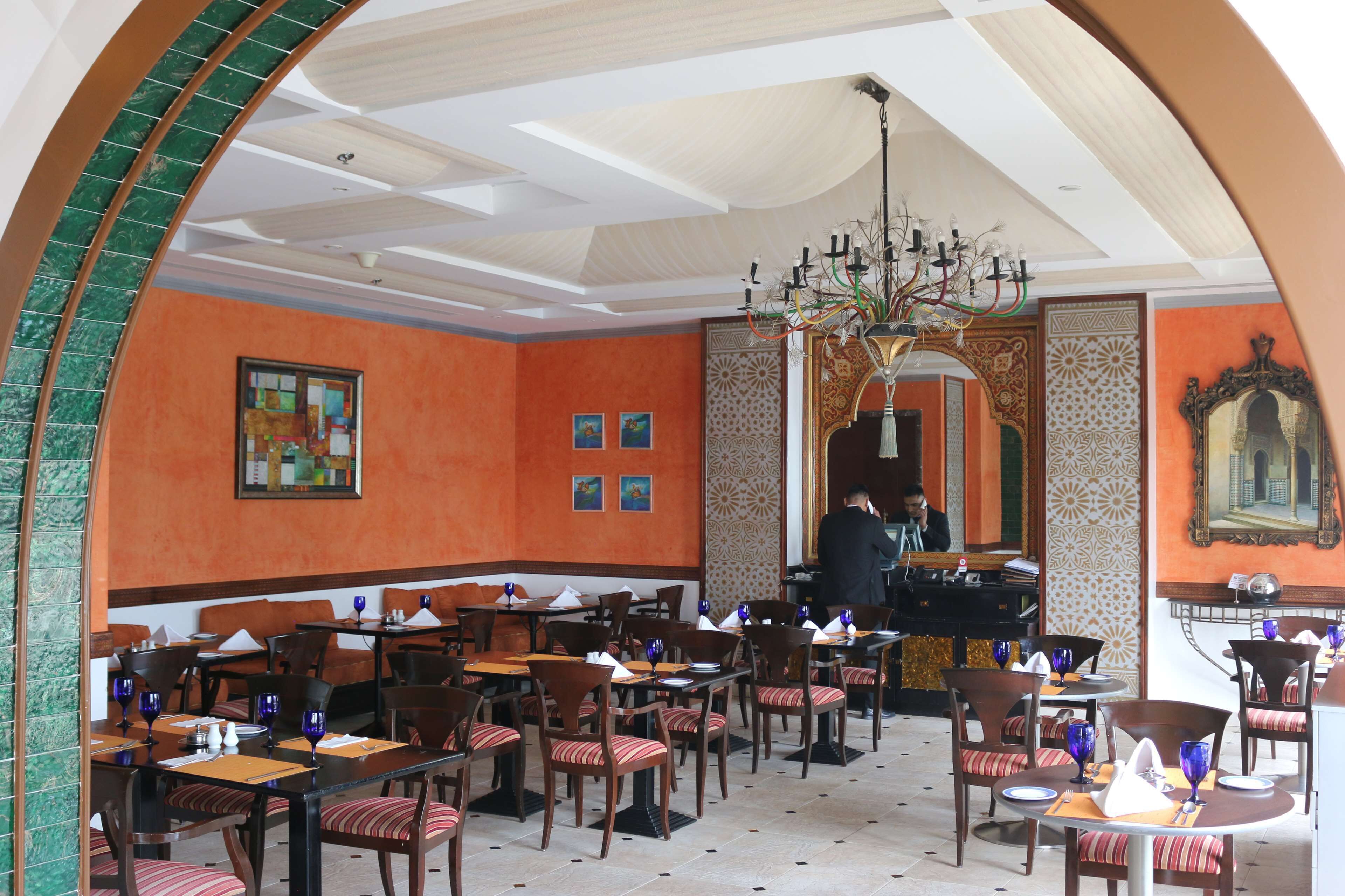 Un restaurante con techo alto, paredes anaranjadas y una araña. Las mesas están preparadas con cristalería azul y servilletas blancas.