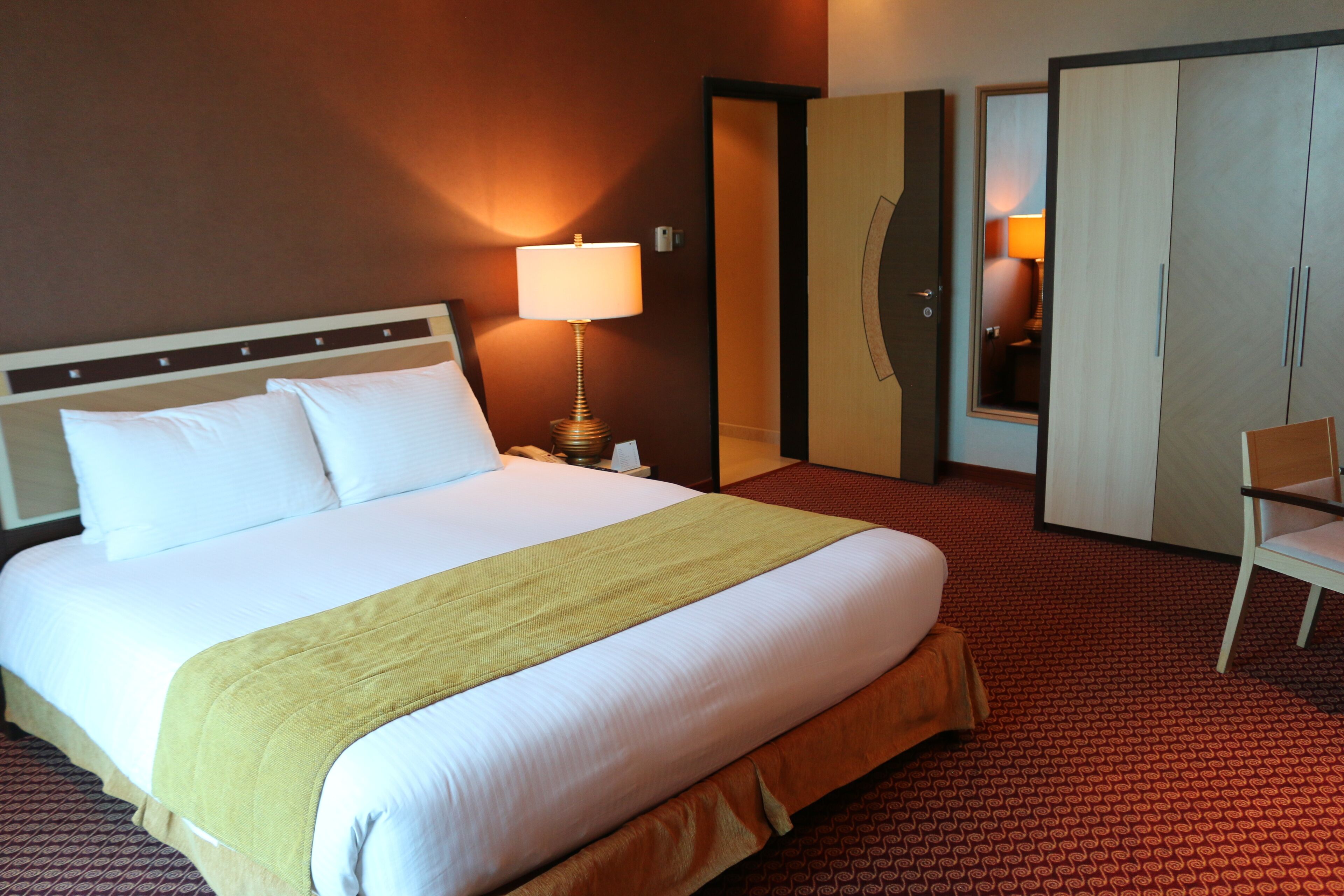 A hotel room with a large bed, a bedside lamp, a wardrobe, and a chair.