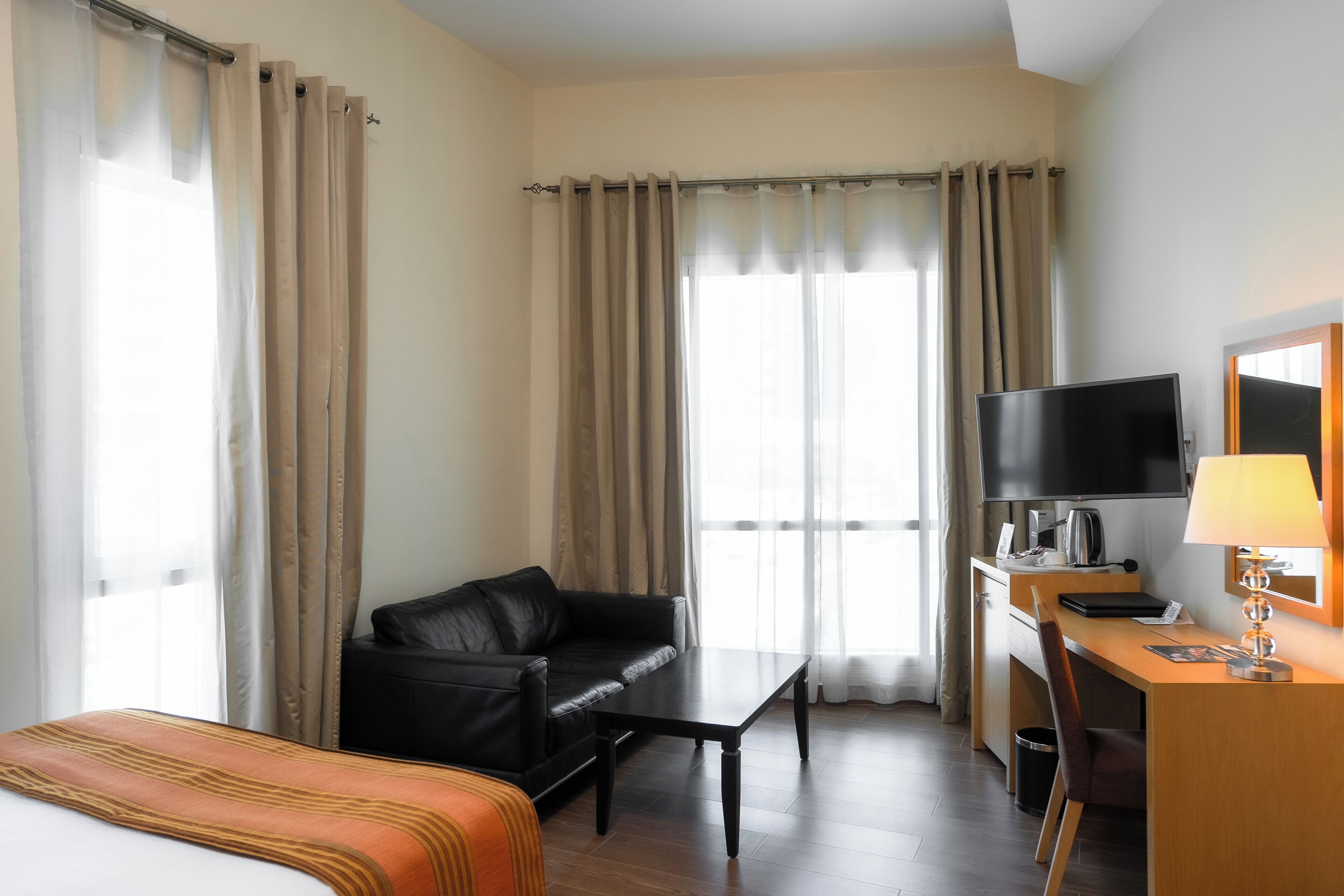 A hotel room with a bed, a black leather sofa, a wooden desk with a television, and a window with curtains.