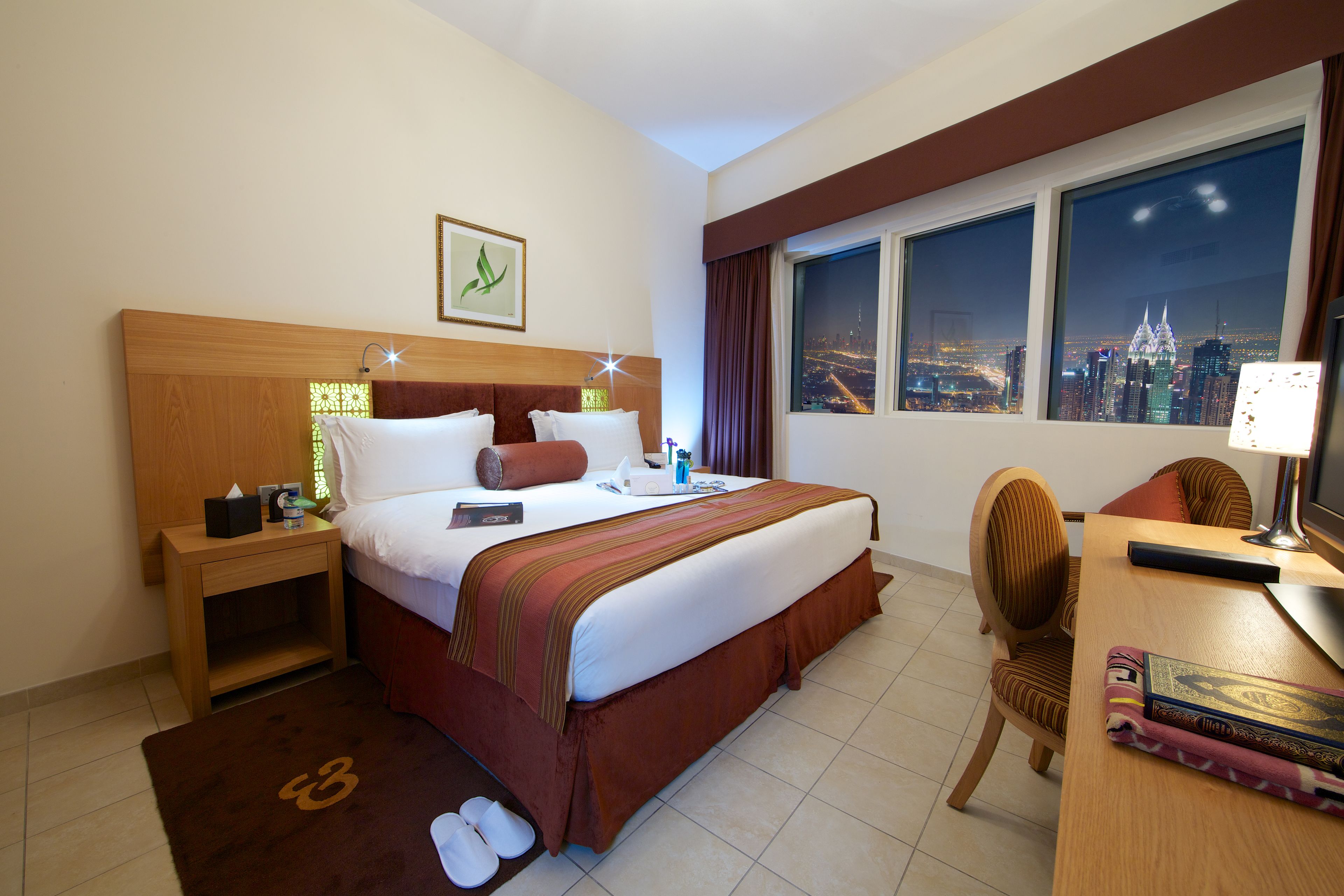 A hotel room with a bed, bedside table, chair, desk, and a view of a cityscape through the window.