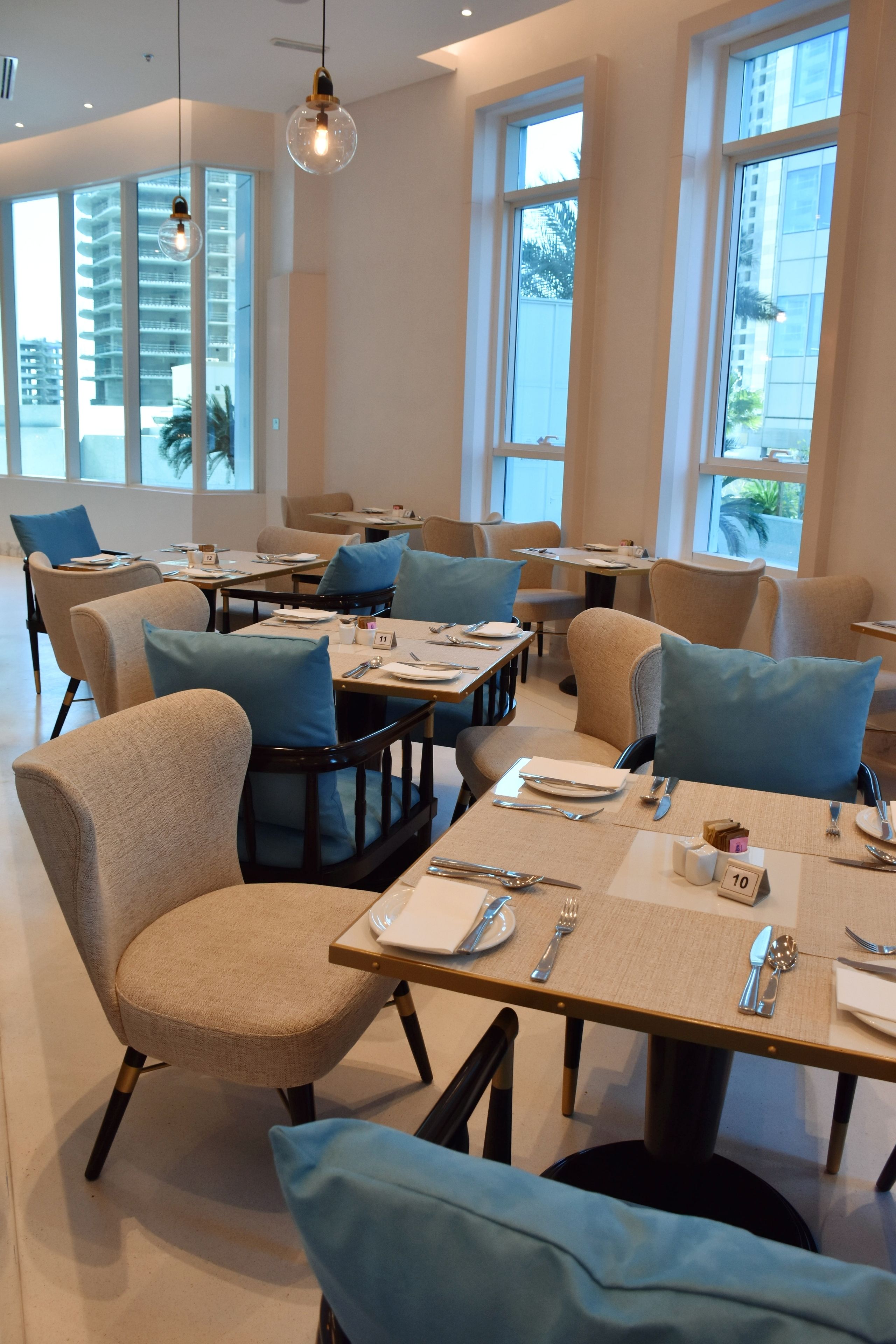 Un comedor moderno con mesas preparadas para dos comensales, con sillas de tonos claros y cojines azules.