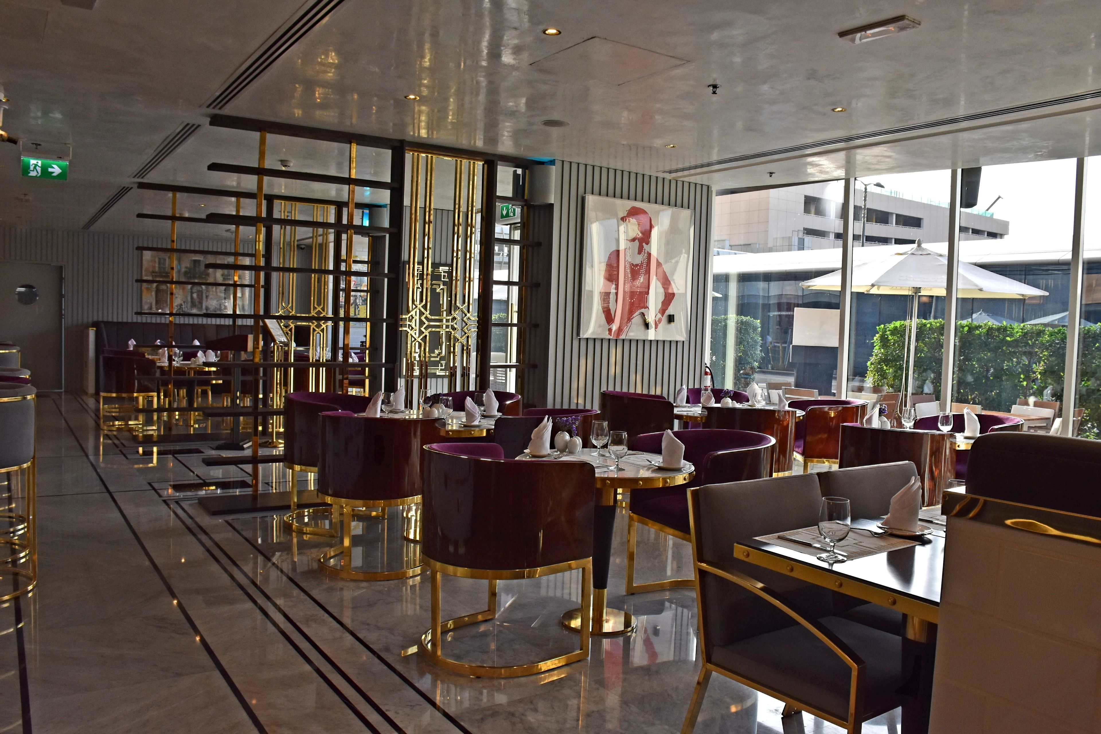 Un restaurante moderno con piso pulido, mobiliario con marcos dorados y grandes ventanales que ofrecen vistas de la ciudad.
