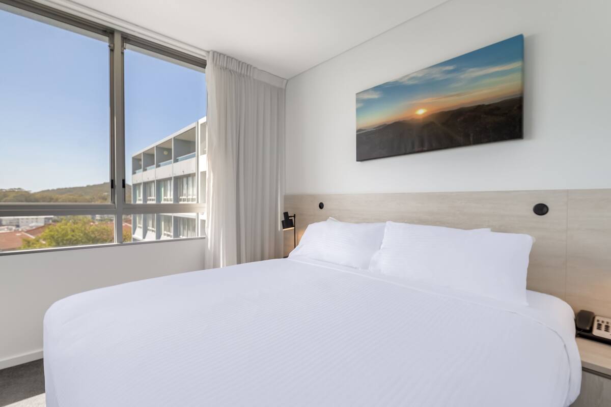 A hotel room with a large bed, a view of the city through a window, and a painting on the wall.