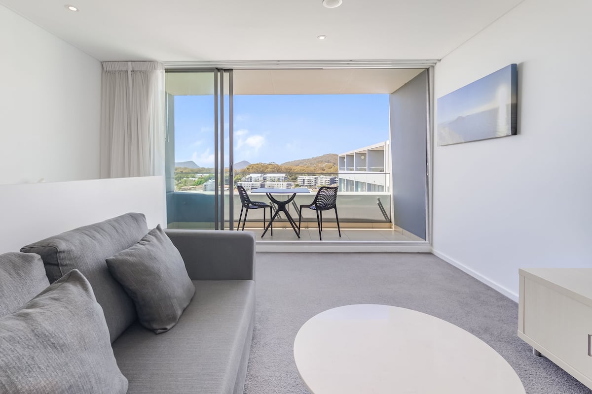 A modern room with a grey sofa, a small white table, and a balcony with a view of buildings and hills.