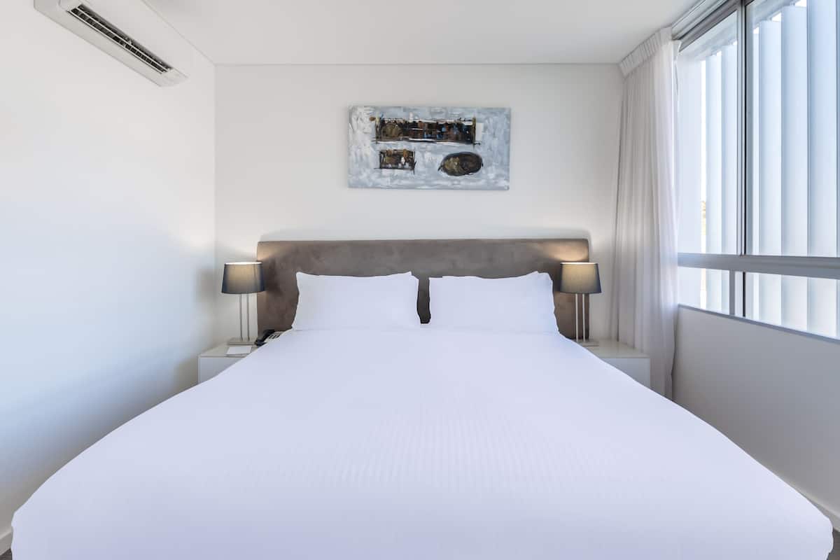 A neatly made bed with white linens, a headboard, bedside lamps, and a wall-mounted artwork.