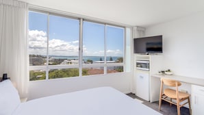 Quarto de hotel com uma cama grande, uma escrivaninha com cadeira, televisão e vista para a cidade e para o mar.