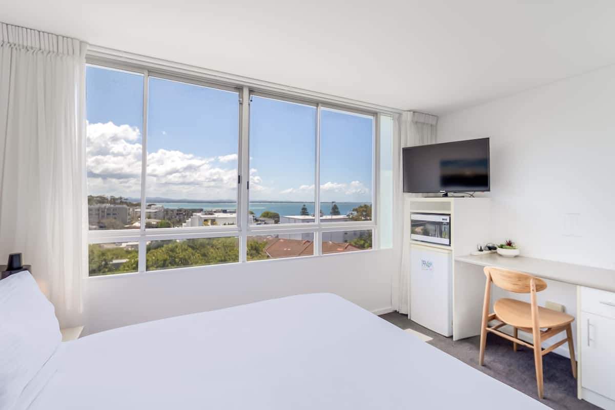 A hotel room with a large bed, a desk with a chair, a television, and a view of the city and sea.
