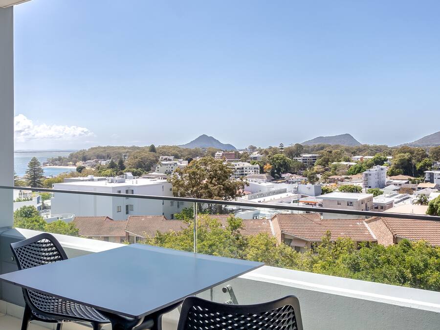 A balcony with a table and chairs, overlooking a coastal town with buildings and mountains in the distance.