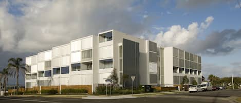 A modern multi-story building with a flat roof, large windows, and a parking area with several cars.