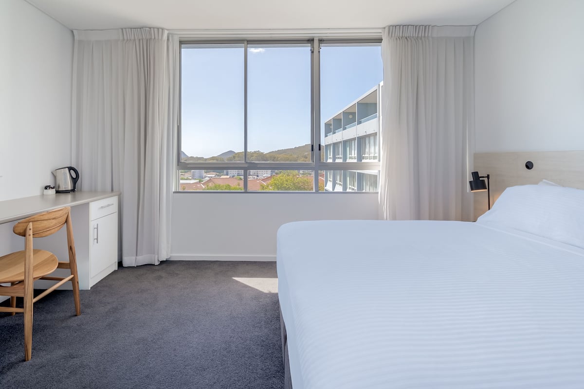 A hotel room with a bed, a desk with a kettle, a chair, and a view of buildings and hills through a large window.