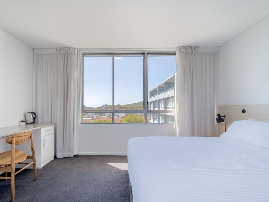 Habitación de hotel con cama, escritorio con tetera, silla y vista a edificios y colinas a través de un ventanal grande.