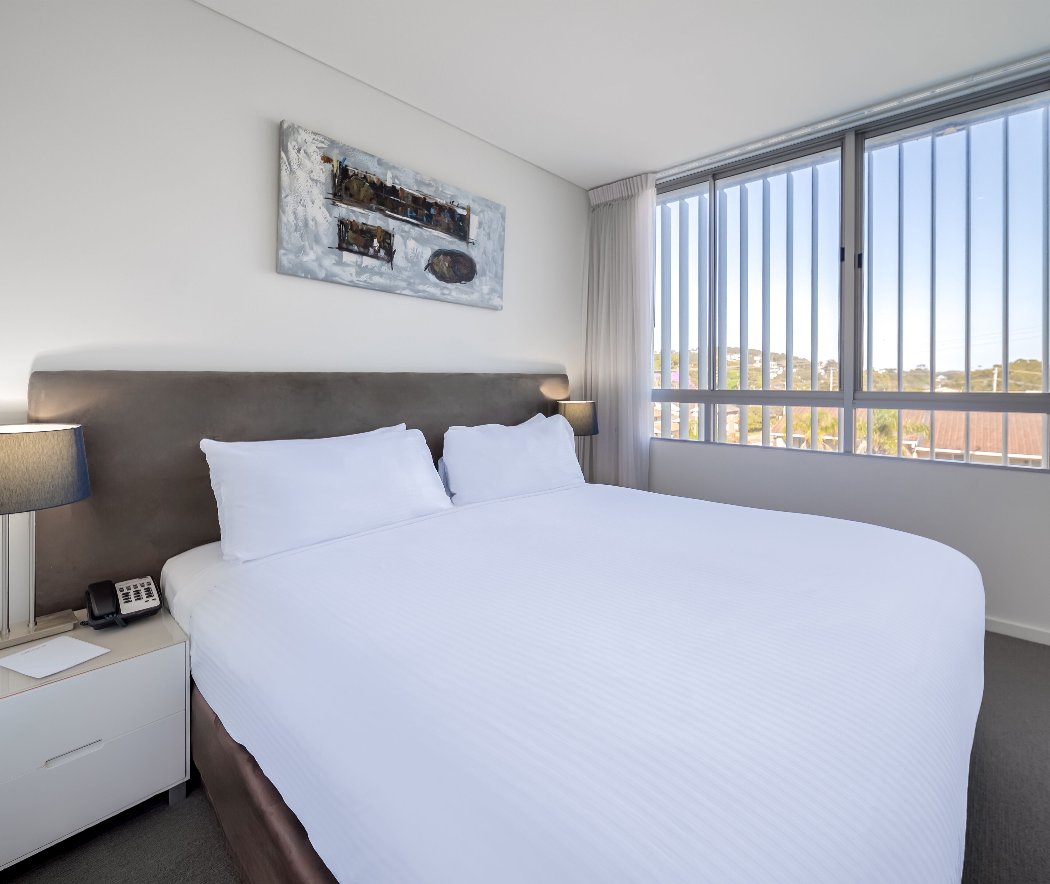 A hotel room with a large bed, a bedside table with a lamp, a phone, and a painting on the wall.