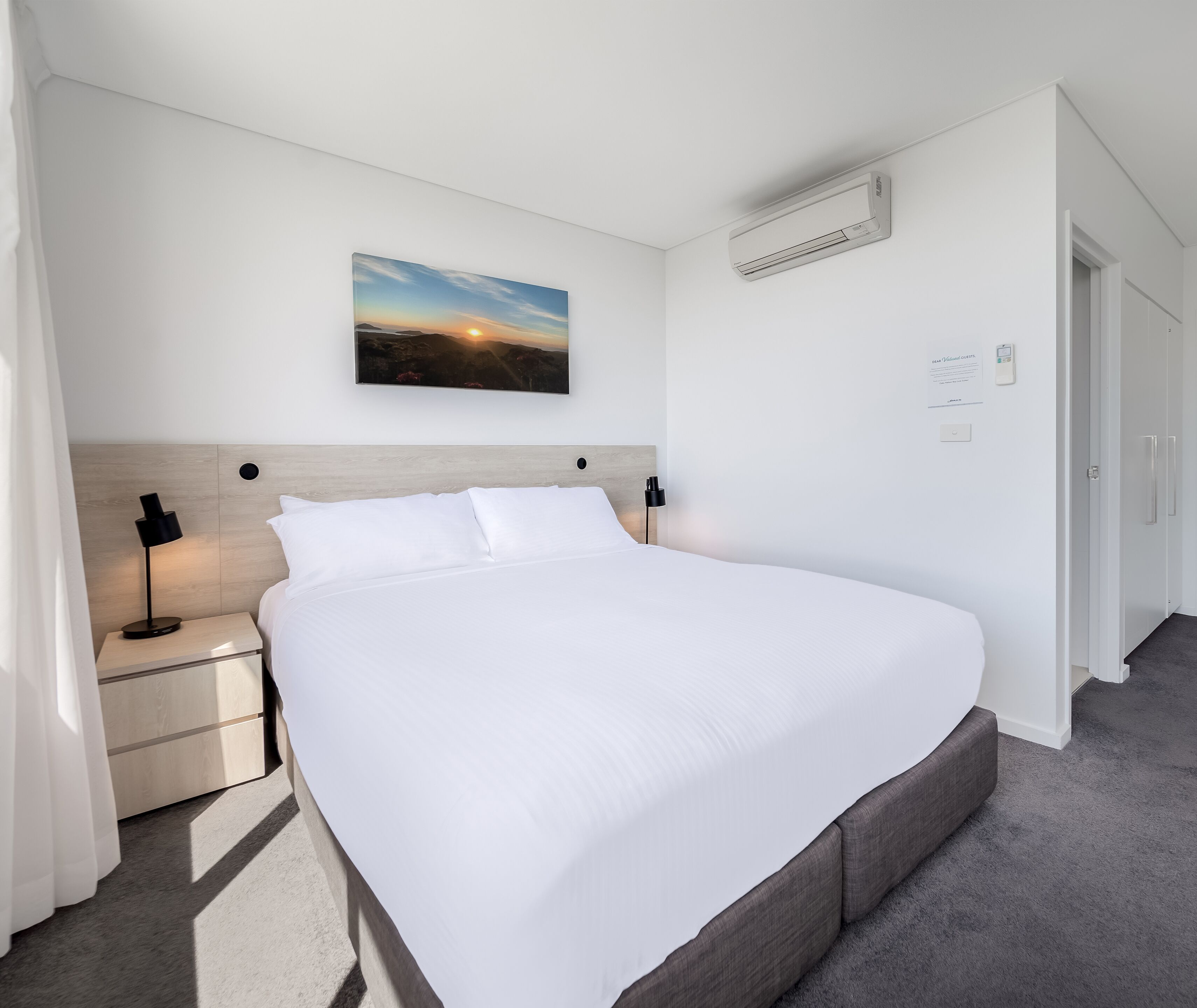 A neatly made bed in a hotel room with a headboard, a bedside table with a lamp, and a painting above the bed.