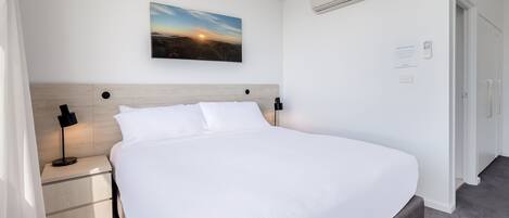 A neatly made bed in a hotel room with a headboard, a bedside table with a lamp, and a painting above the bed.