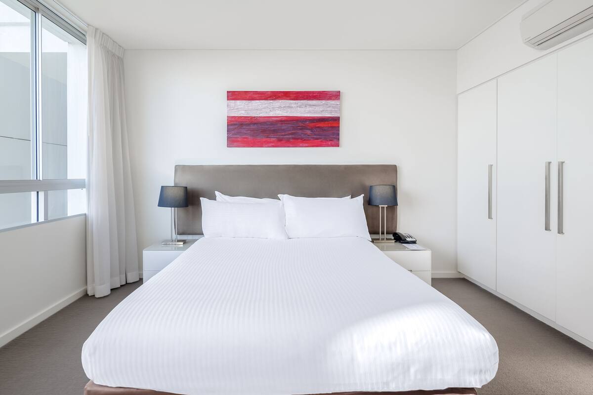 A modern hotel room with a large bed, a nightstand with a lamp, a telephone, and a wall-mounted artwork.