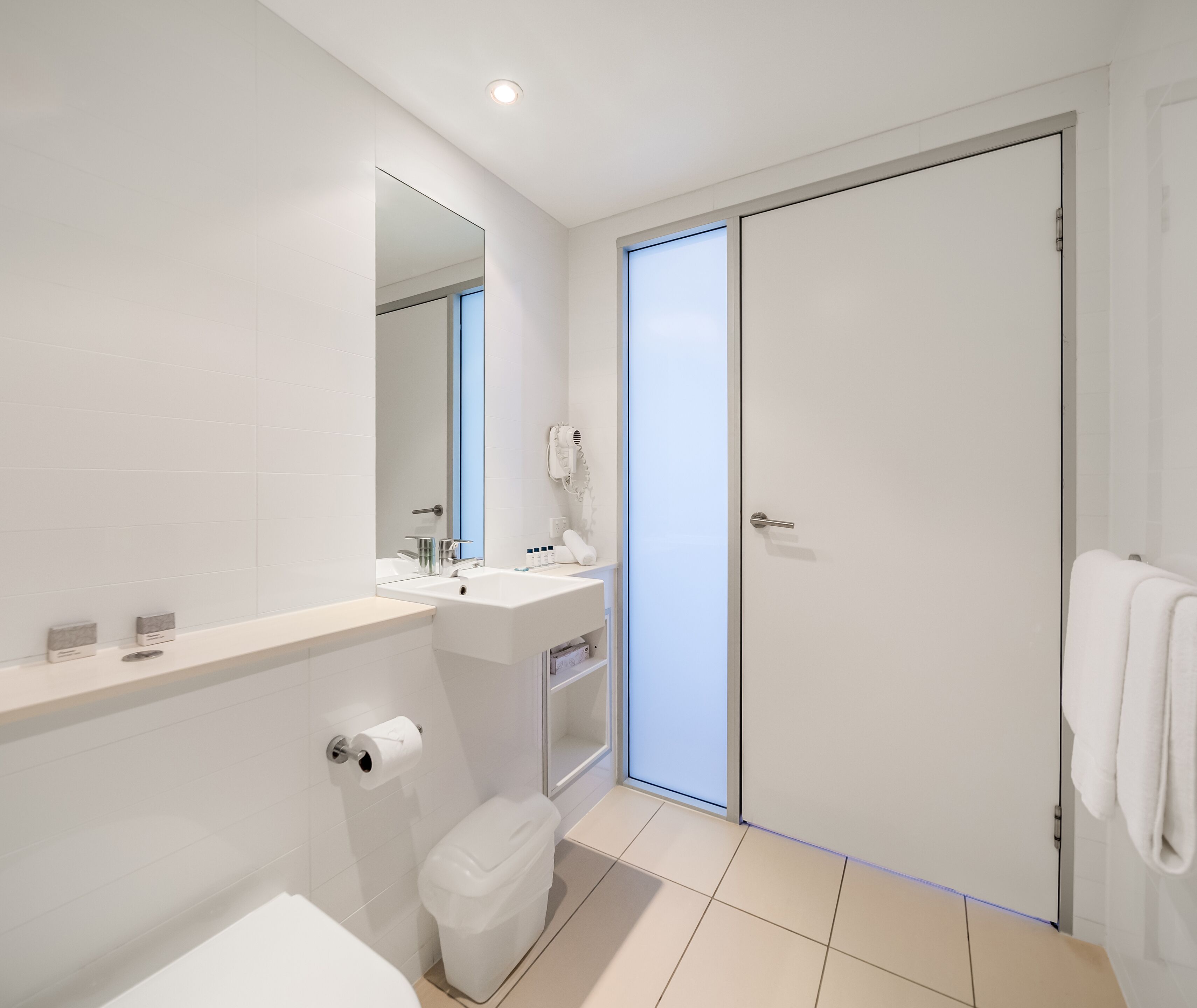 A modern bathroom with a white sink, mirror, and toilet. There is a towel rack and a shower area visible.
