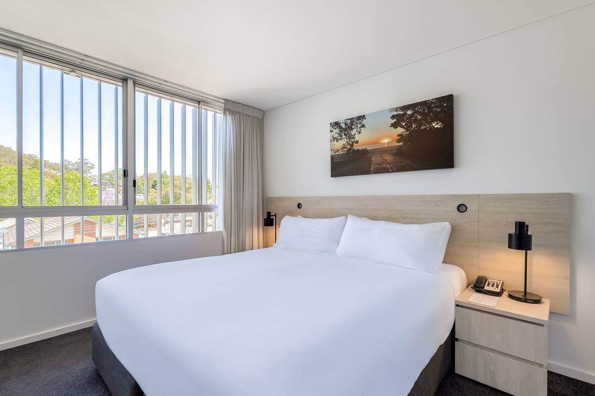A hotel room with a large bed, a bedside table with a lamp, a phone, and a painting on the wall.