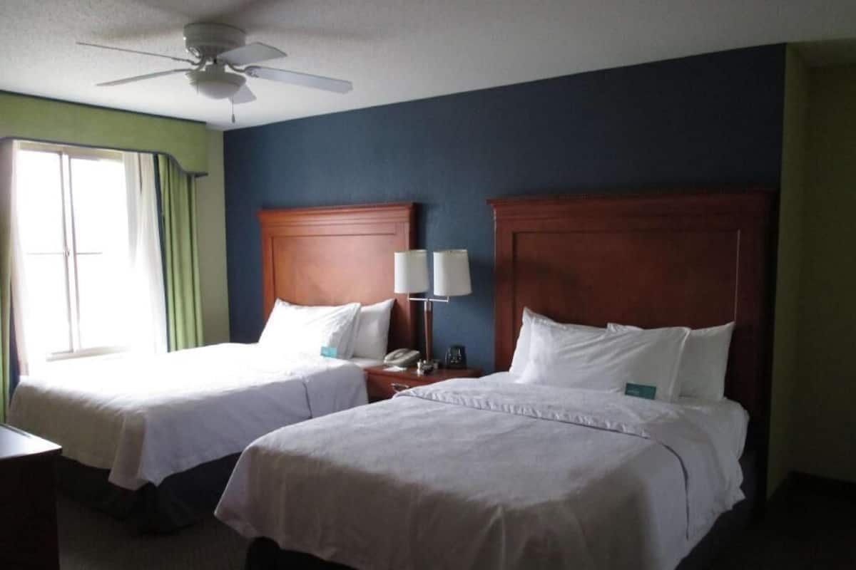 Two single beds with wooden headboards, a ceiling fan, a bedside table with a lamp, and a window with curtains.