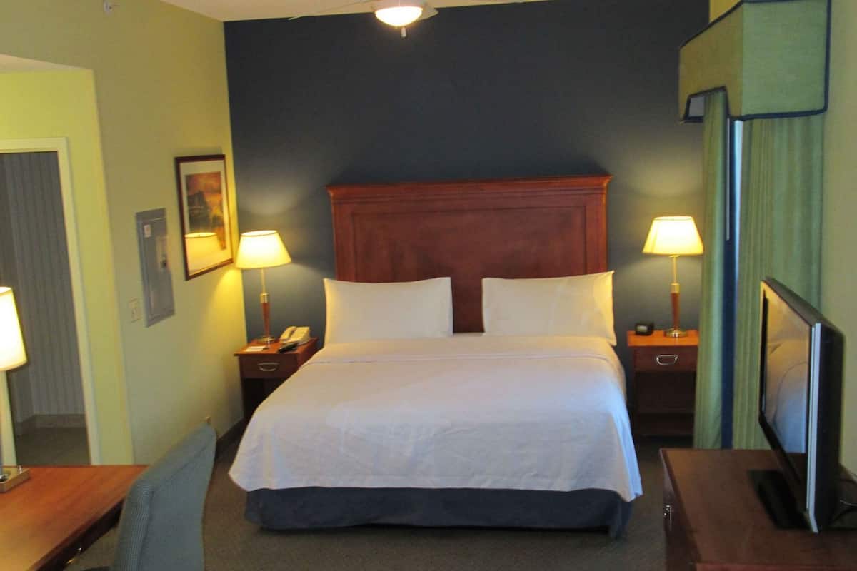 A hotel room with a bed, two bedside lamps, a desk with a chair, a television, and a ceiling fan.