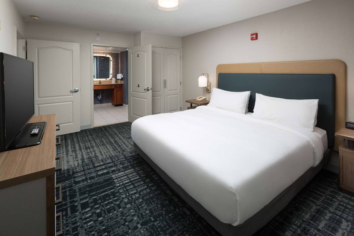 A hotel room with a large bed, a desk with a computer, a telephone, and a bathroom visible through an open door.