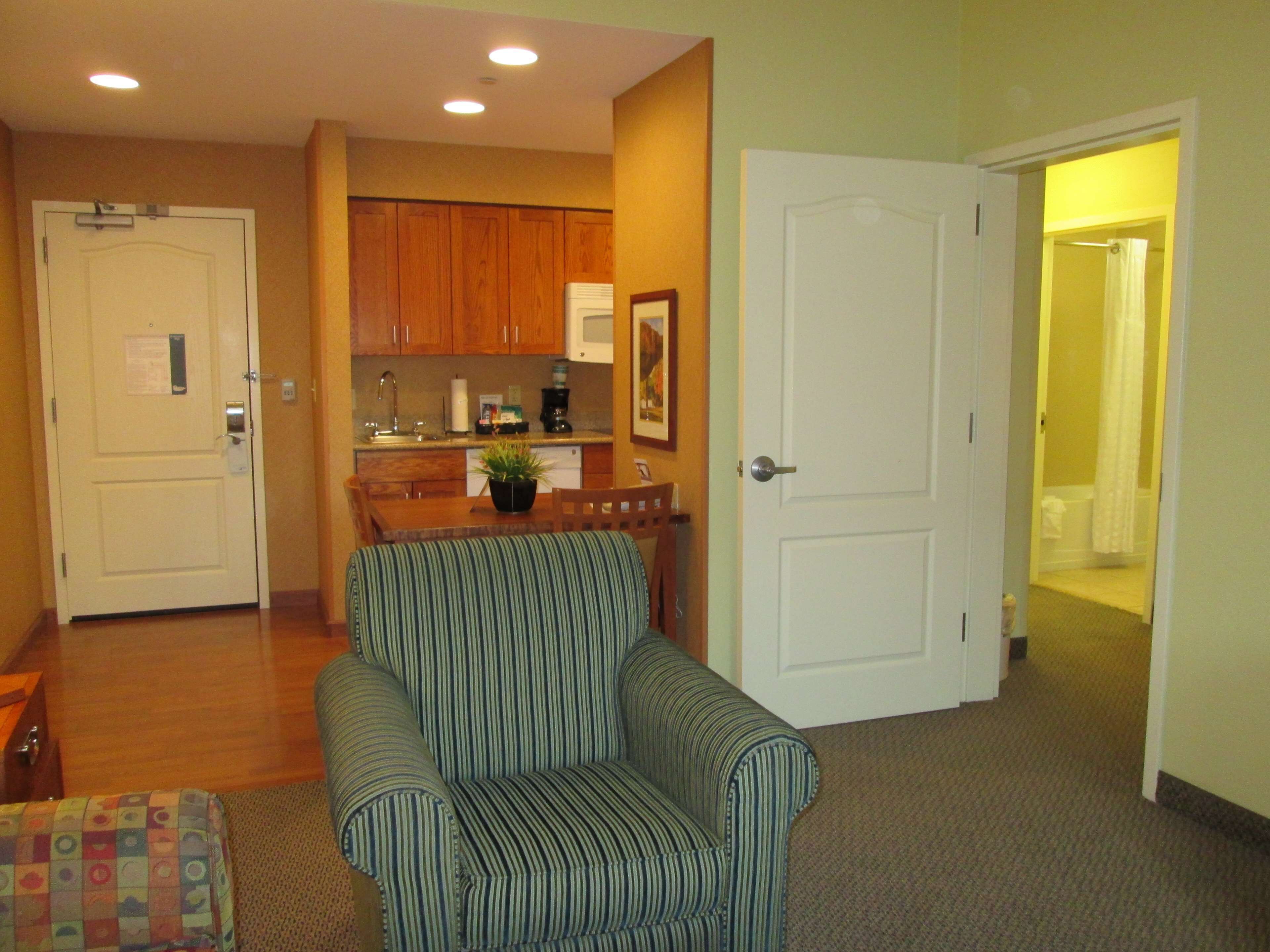 A living room with a striped armchair, a small table with a plant, and a kitchen area with wooden cabinets and a microwave.