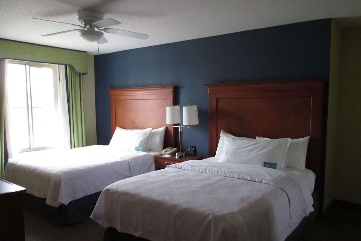 A hotel room with two beds, a ceiling fan, a nightstand with a lamp, and a window with curtains.