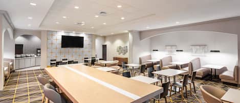 A modern hotel lobby with a long wooden reception desk, multiple seating areas, and a flat-screen TV mounted on the wall.