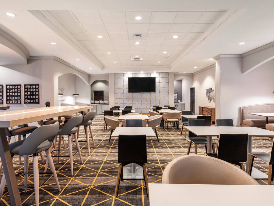 A modern hotel lobby with a central bar, multiple seating areas, and a large flat-screen TV on the wall.