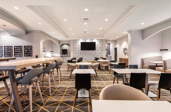 Een moderne hotellobby met een centrale bar, meerdere zithoeken en een grote flatscreen televisie aan de muur.