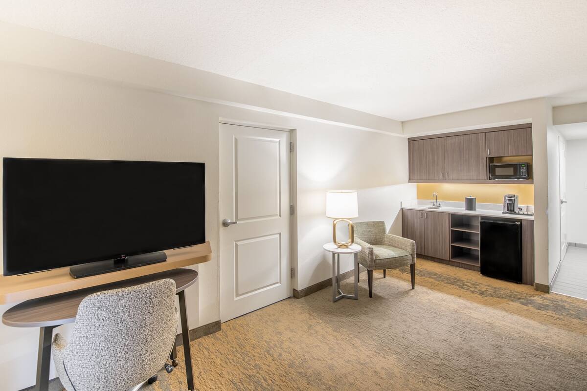 A hotel room with a flat-screen TV, a small table with a lamp, a chair, a kitchenette with a microwave, and a refrigerator.