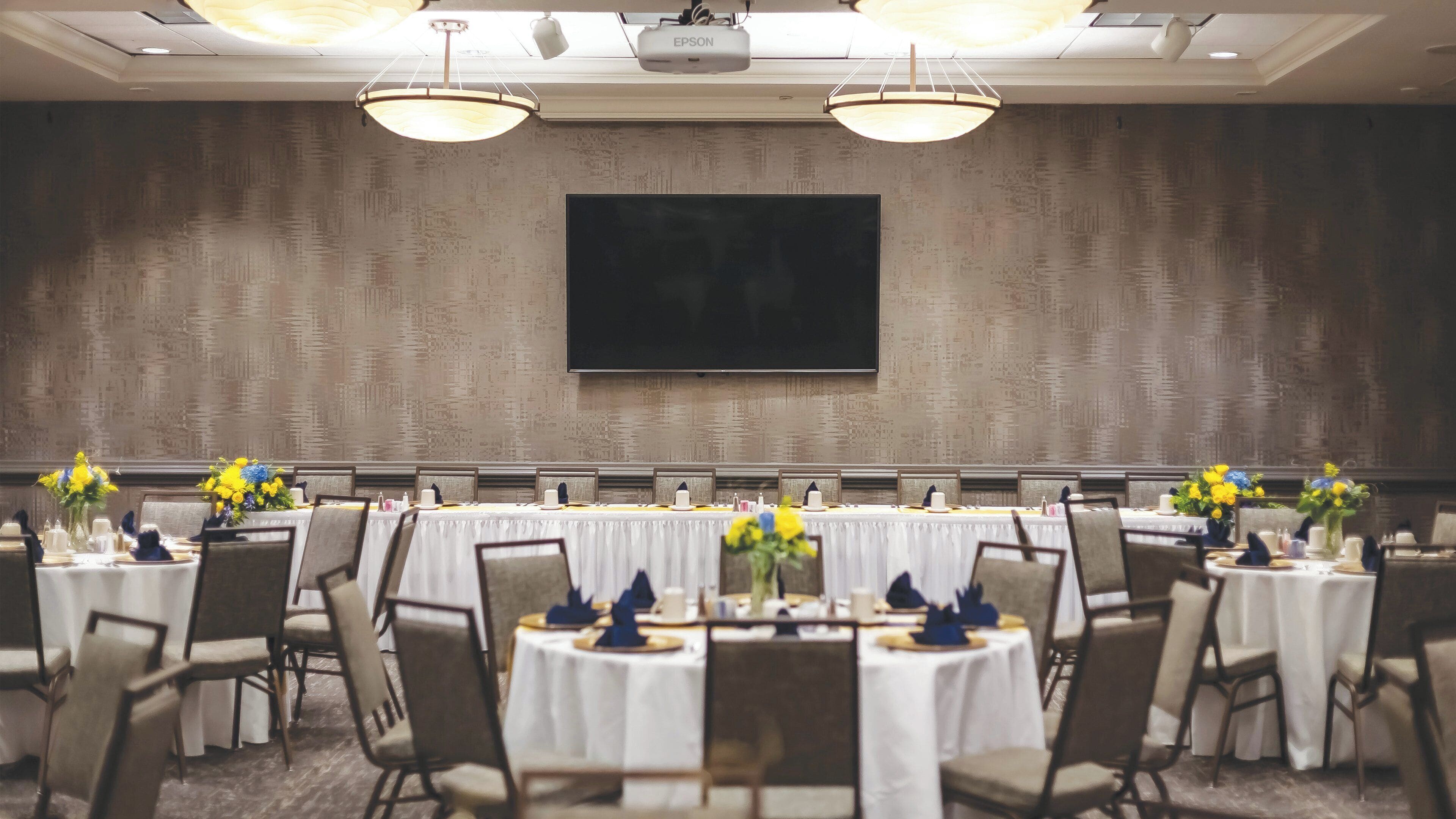 Un salón de conferencias con una televisión de pantalla plana grande montada en la pared, mesas redondas con manteles blancos, sillas y centros de mesa con flores.