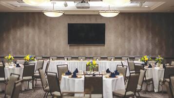 Un salón de conferencias con una televisión de pantalla plana grande montada en la pared, mesas redondas con manteles blancos, sillas y centros de mesa con flores.