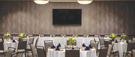 A conference room with a large flat-screen TV mounted on the wall, round tables with white tablecloths, chairs, and flower centerpieces.