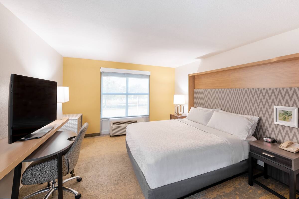 A hotel room with a bed, desk, chair, TV, bedside table, lamp, and a window.
