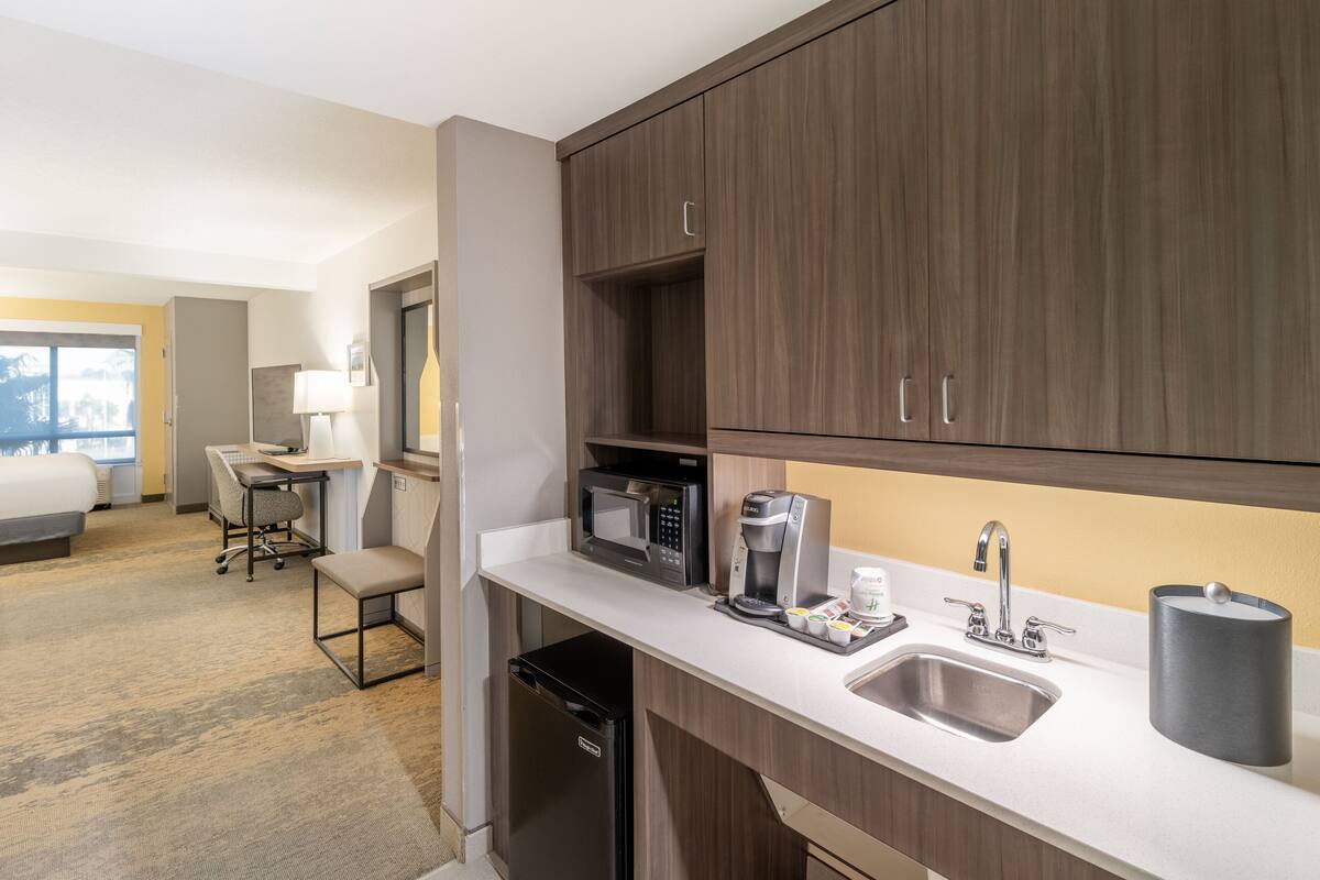 A hotel kitchen with a microwave, coffee maker, sink, and a small dining area with a chair and table.