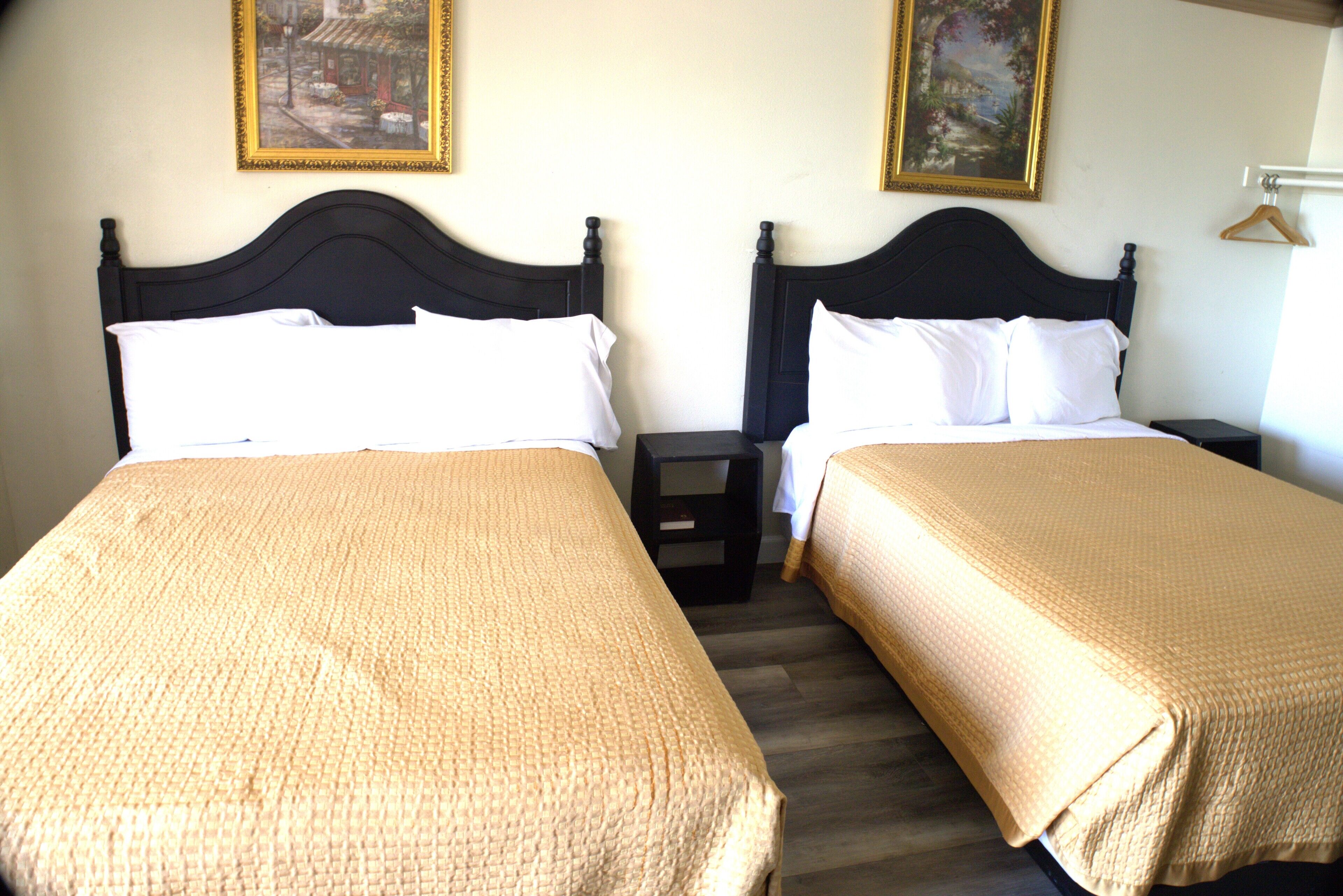 A hotel room with two beds, each with a dark headboard and white bedding. There are two paintings on the walls, a small nightstand between the beds, and a wooden hanger on the wall.