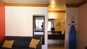 A room with a patterned sofa, a blue ironing board, and a kitchen area visible through an open doorway. - Lambert Inn (Whittier)