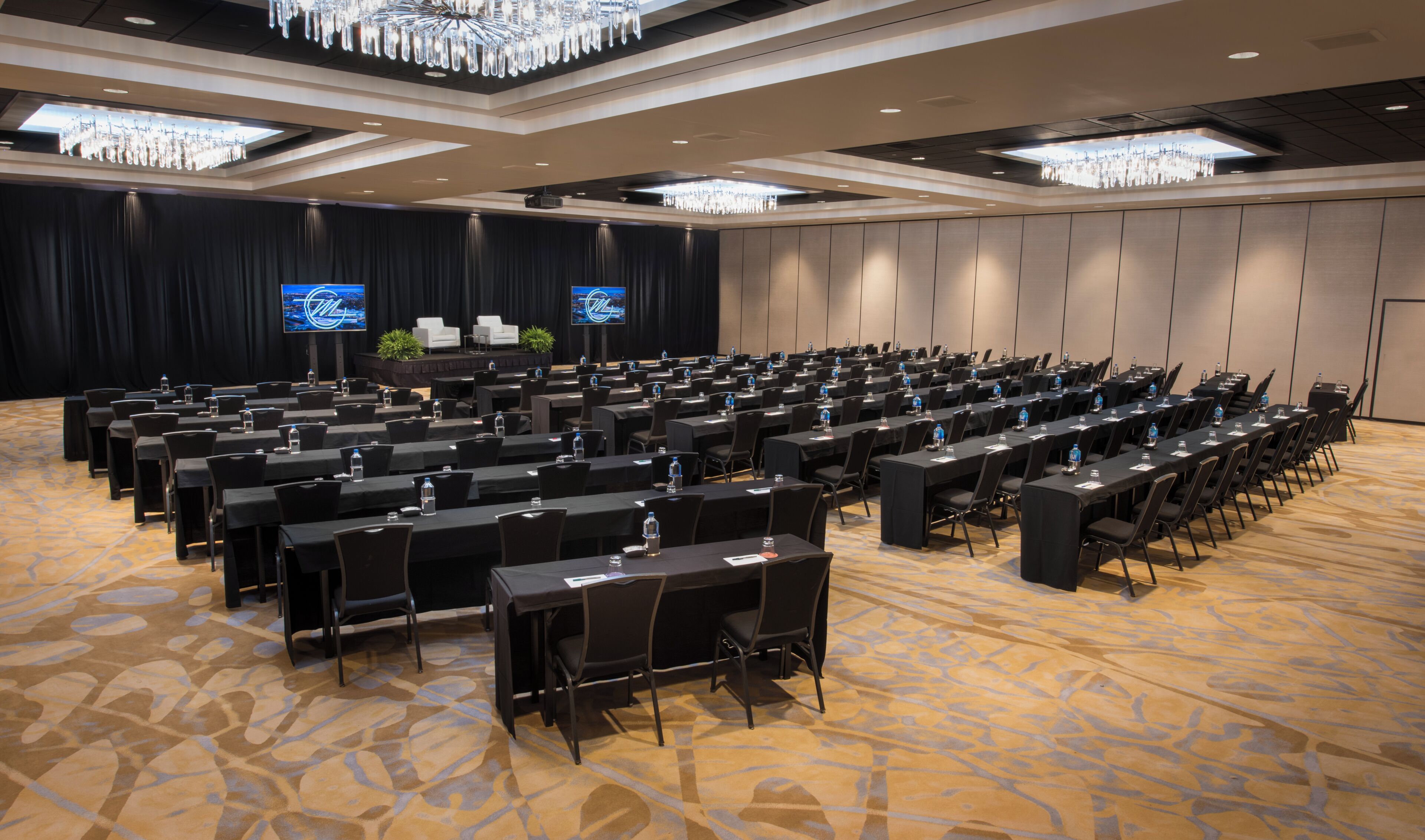 A conference room with rows of tables and chairs, a stage with a screen, and a chandelier.