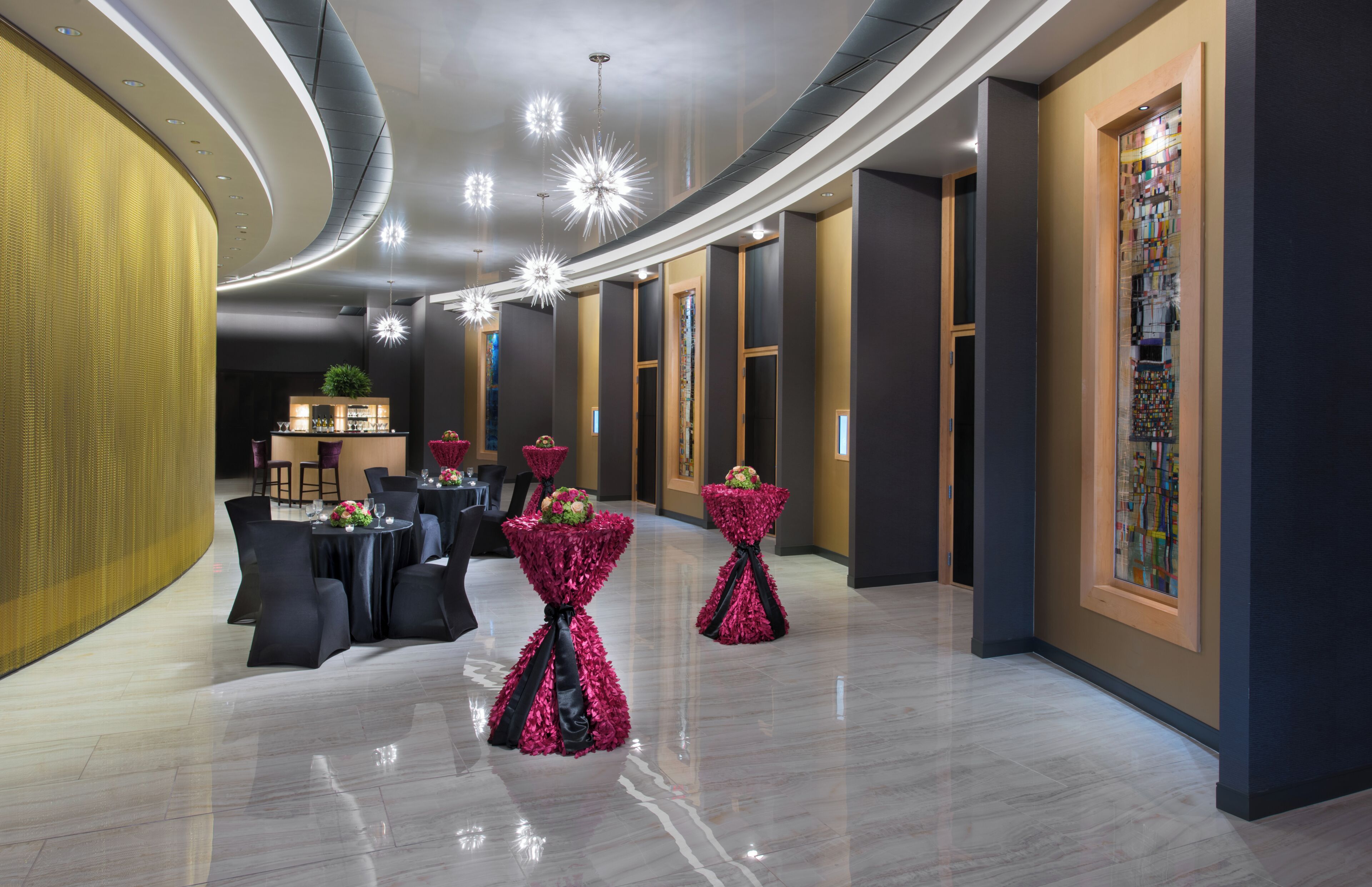 A modern hotel lobby with a bar area, round tables, and decorative floral arrangements.