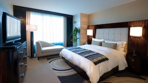 Ein Hotelzimmer mit einem großen Bett, einem Schreibtisch mit Flachbildfernseher, einem Sessel, einer Pflanze und einem Nachttisch mit Lampe.